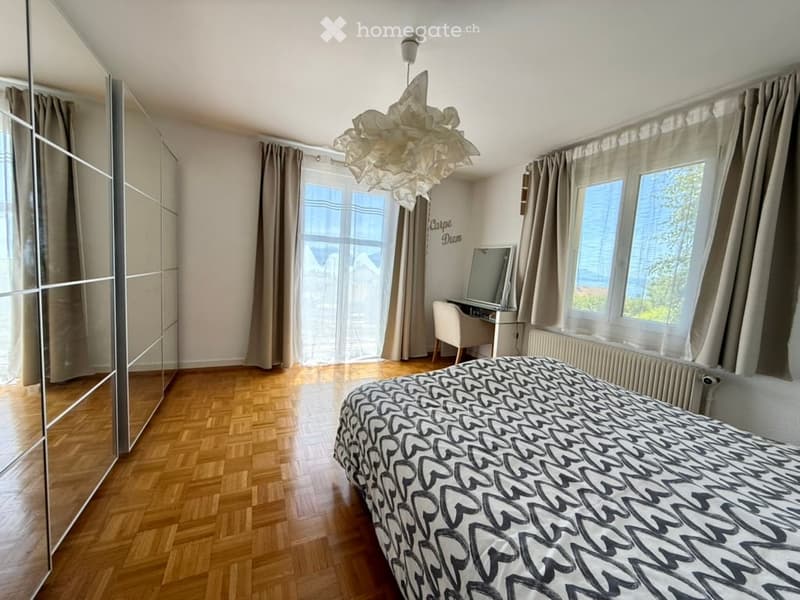 Bright bedroom with wooden flooring, large windows offering a view, a bed with patterned bedding, a wardrobe, a desk with a chair, and a decorative hanging lamp.