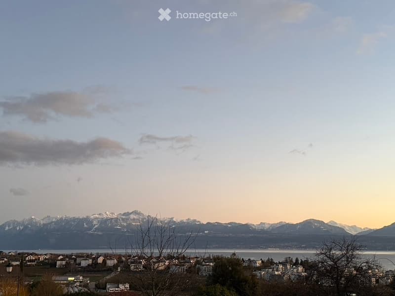 Panoramic view of a town with houses, surrounded by mountains and a body of water under a partly cloudy sky.