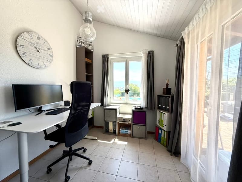 Bright, modern home office with a large desk, ergonomic chair, computer setup, and large windows offering a scenic view. Includes a clock, storage cabinets, and decorative items.