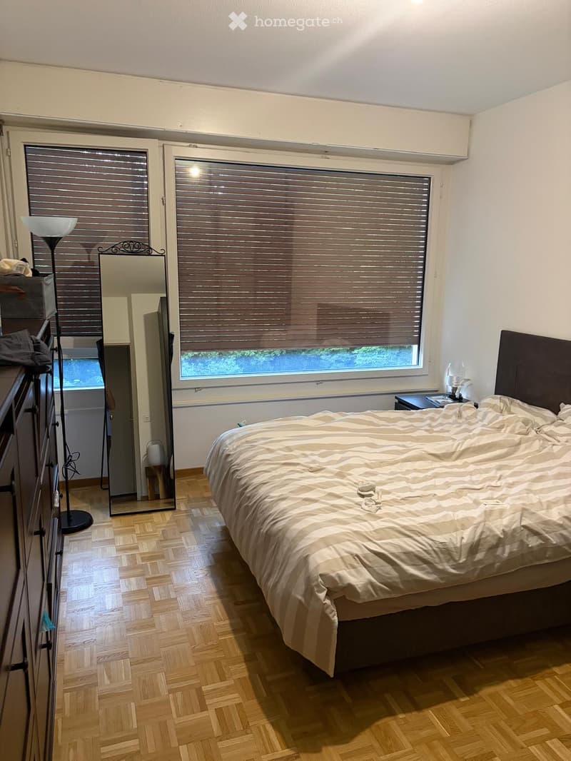 Cozy bedroom with wooden flooring, large window with blinds, neatly made bed, dresser, mirror, and floor lamp.