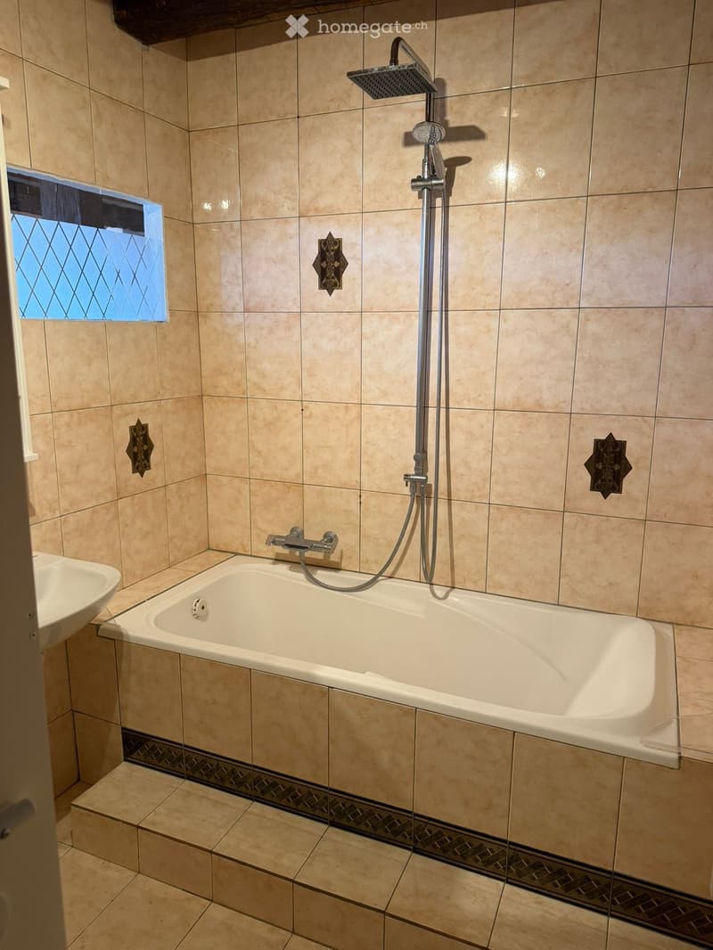 Bathroom with beige tiled walls and floor, a bathtub with a handheld showerhead, a sink, and decorative wall tiles.