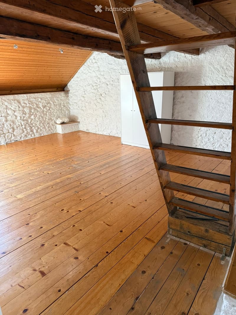 An attic space with wooden floors and exposed wooden beams, featuring a staircase leading upwards and a built-in shelf on the wall.