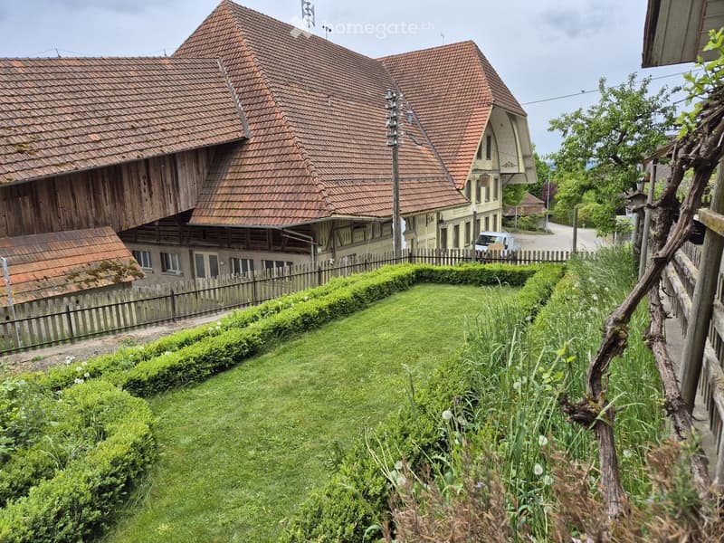 Traditional wooden house with red-tiled roof, surrounded by a well-maintained garden with hedges and flowers, and a wooden fence.