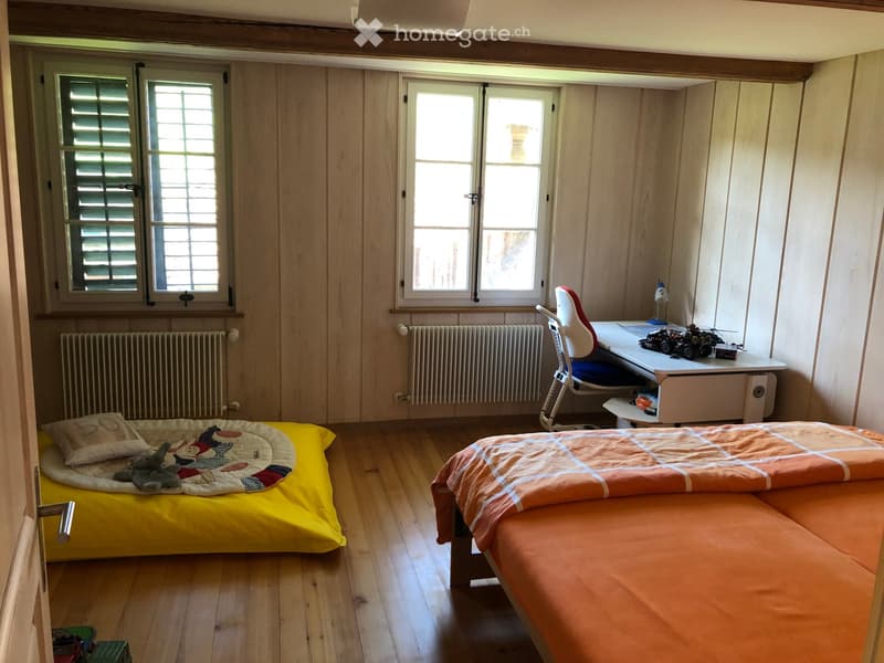 Cozy bedroom with wooden walls and flooring, featuring a double bed with orange bedding, a daybed with yellow bedding, a desk with a chair, and a small toy car on the desk.