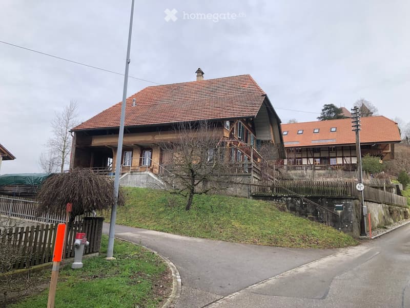 Two-story house with red-tiled roof, wooden balcony, and stone foundation, situated on a hillside with a wooden fence and small garden area.