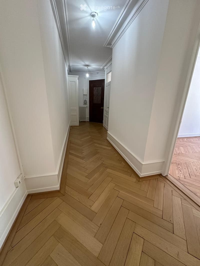 A long, narrow hallway with herringbone-patterned wooden flooring, white walls, and recessed lighting. A closed door is visible at the end of the hallway, along with a radiator and a small cabinet on the left side.