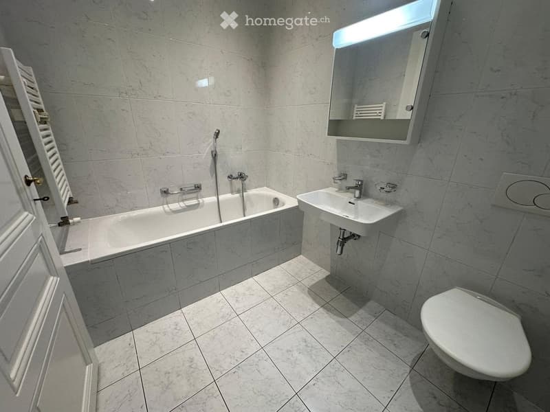 Modern bathroom with marble tiles, a bathtub, a sink, a toilet, and a mirror with built-in lighting.