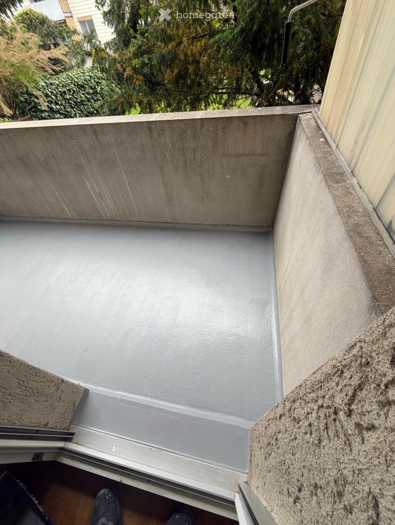 A small, enclosed balcony with a concrete floor and walls, featuring a metal safety railing and a sliding glass door leading to the interior.
