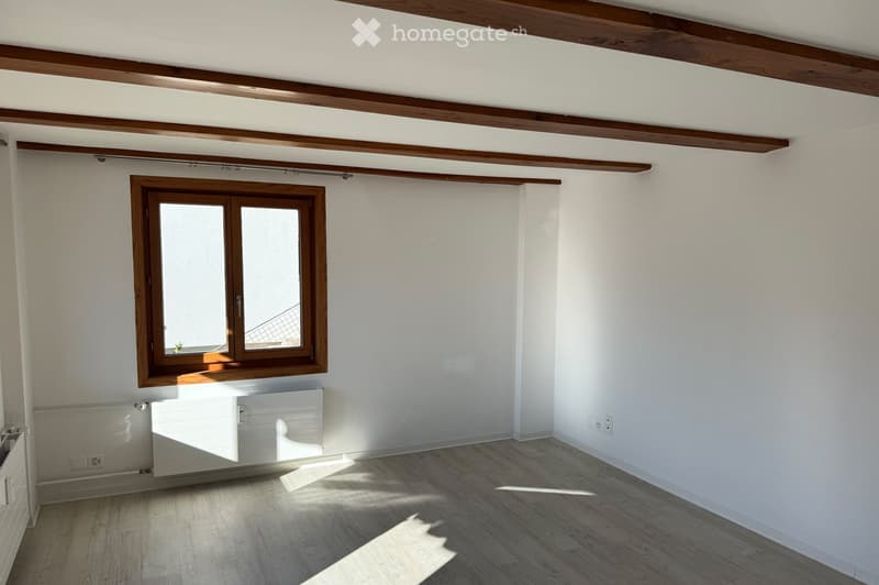 Empty room with wooden floor, white walls, exposed wooden beams on the ceiling, and a single window with wooden frame. No furniture or equipment present.