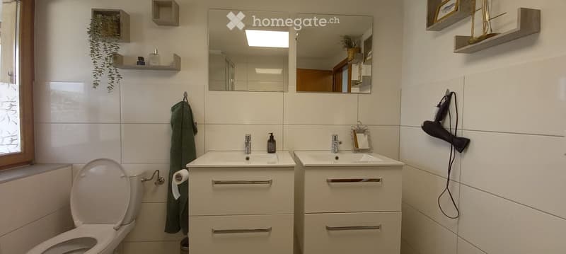 Modern bathroom with white tiles, two sinks, a toilet, a mirror, a hair dryer, and decorative shelves.