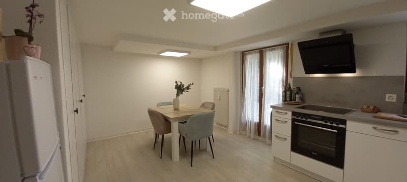 Modern kitchen and dining area with white cabinets, stainless steel appliances, a dining table with chairs, and a refrigerator. The space is well-lit with natural light from a window with white curtains.