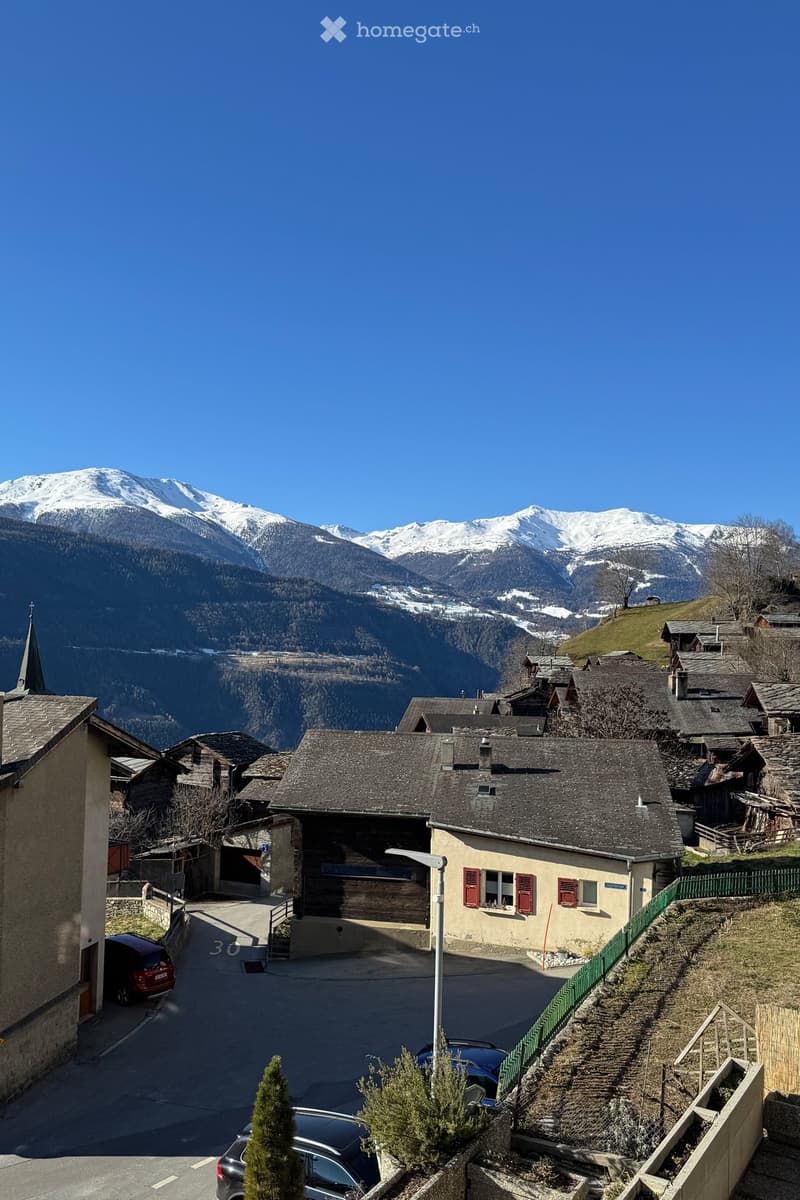 A picturesque village scene with traditional stone houses, red shutters, and a snow-capped mountain backdrop. The village features narrow streets, parked cars, and a small green fence along a hillside.