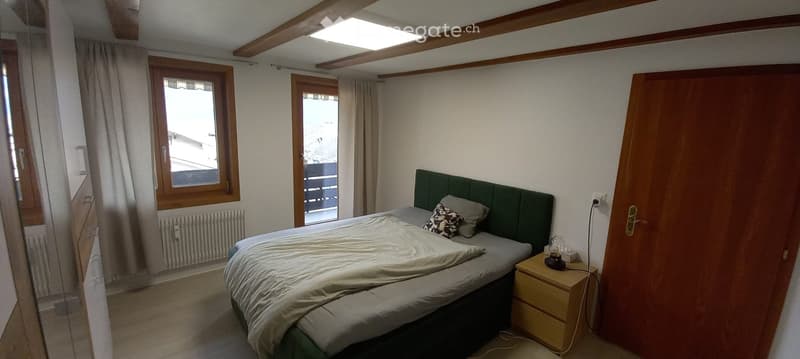 Cozy bedroom with a double bed, nightstand, and large windows with curtains. The room features exposed wooden beams on the ceiling and a radiator under one window.