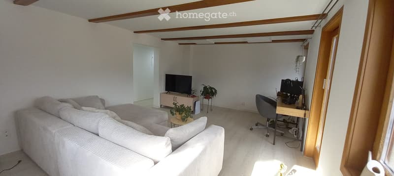 Bright living room with a white sofa, ottoman, coffee table, and a small desk with a computer. The room features wooden beams on the ceiling and a large window letting in natural light.