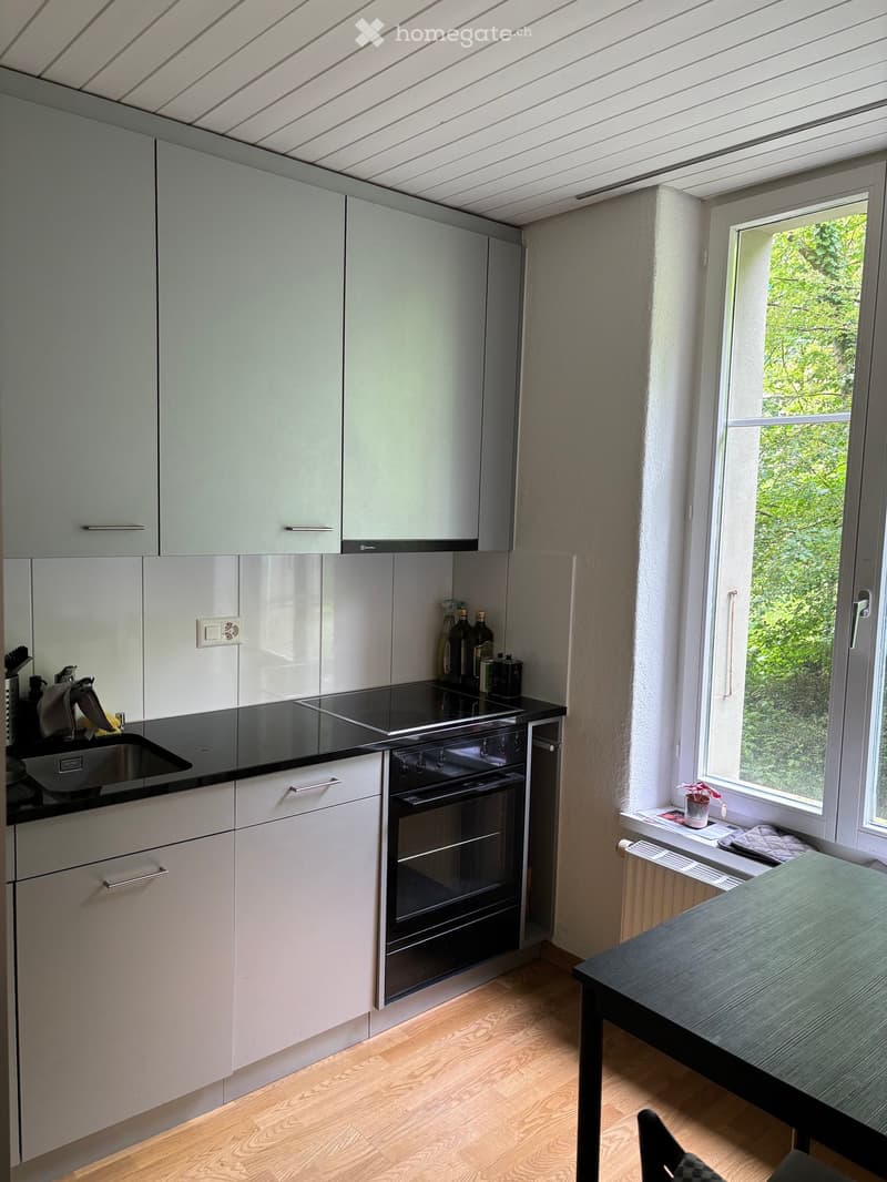 Modern kitchen with light grey cabinets, black countertops, and stainless steel appliances including a sink, faucet, and oven. A small table and various bottles are visible.