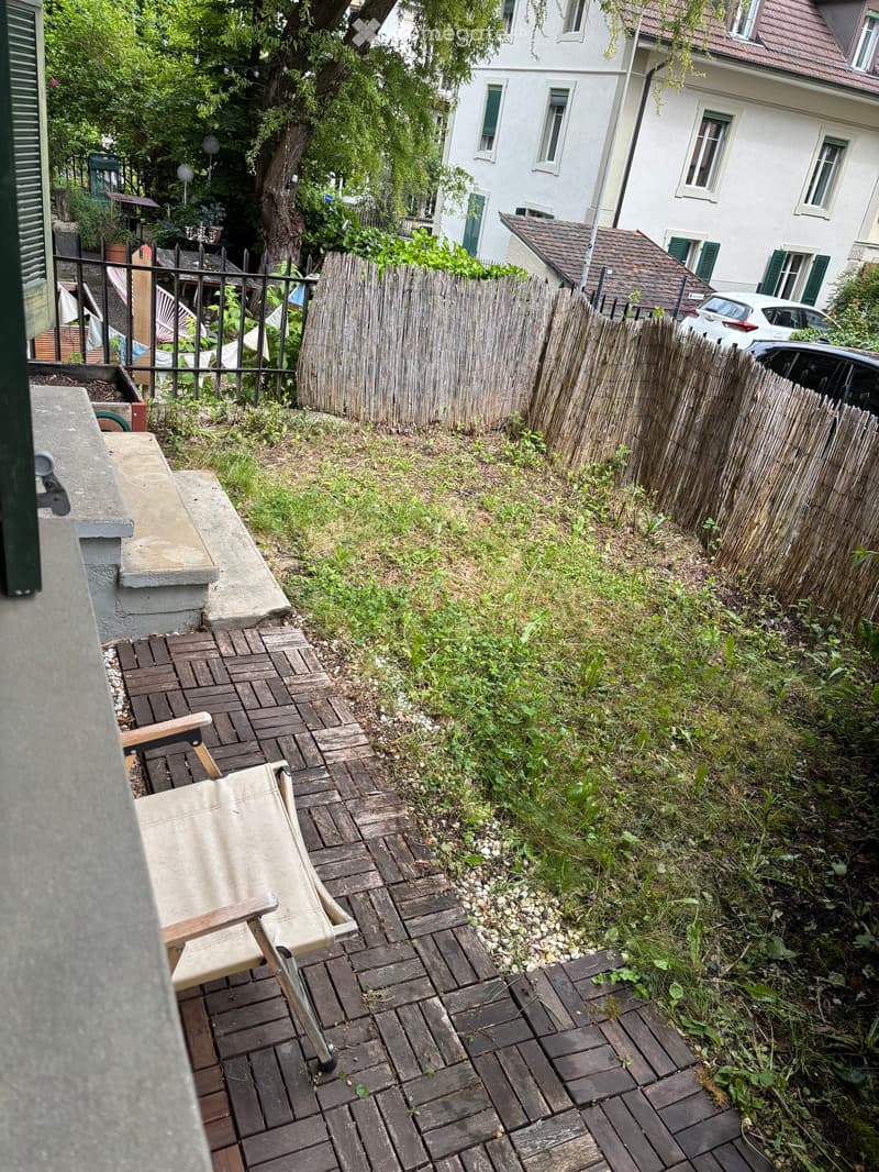 Small outdoor area with brick patio, wooden chair, grassy slope, and wooden fence.