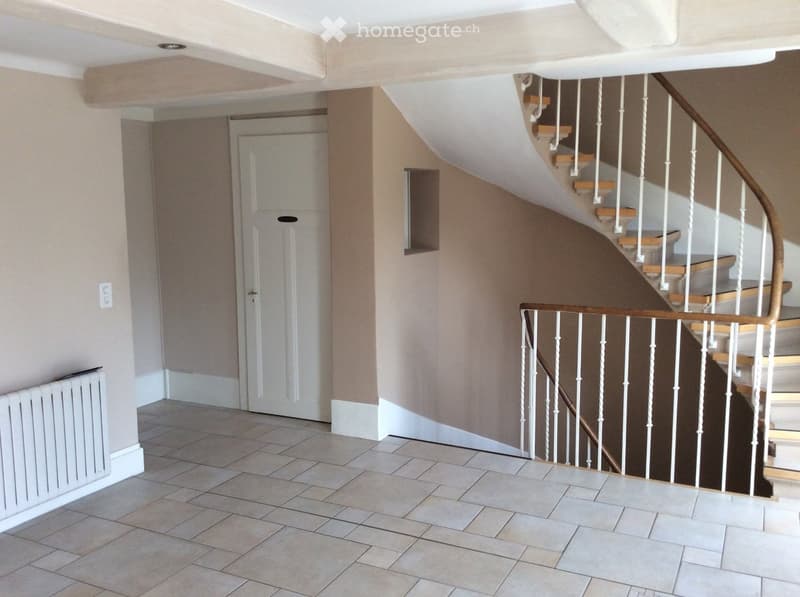 Spacious hallway with tiled flooring, white walls, a staircase with wooden steps and white balustrade, and a closed door leading to another room.