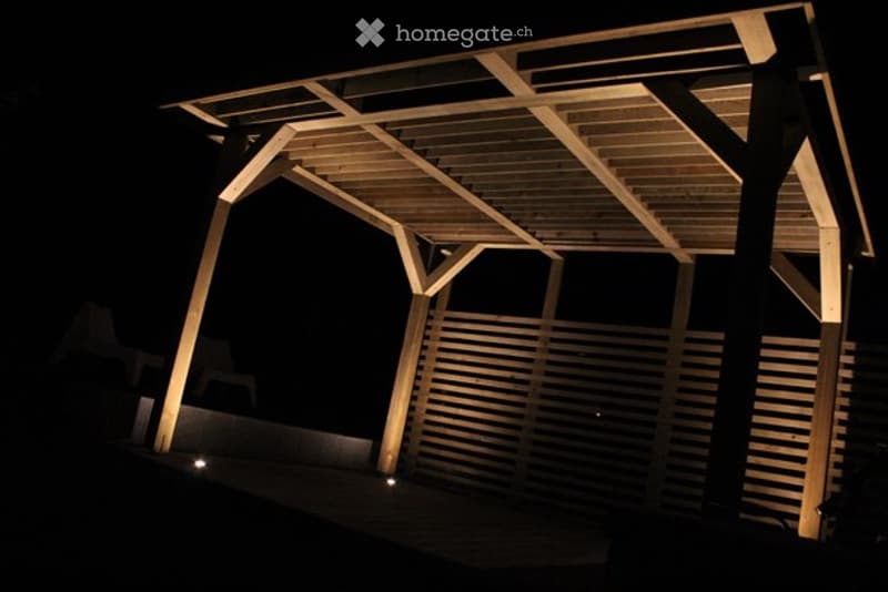 A wooden pergola structure illuminated by warm lighting, featuring a slatted roof and vertical wooden panels, set against a dark night sky.