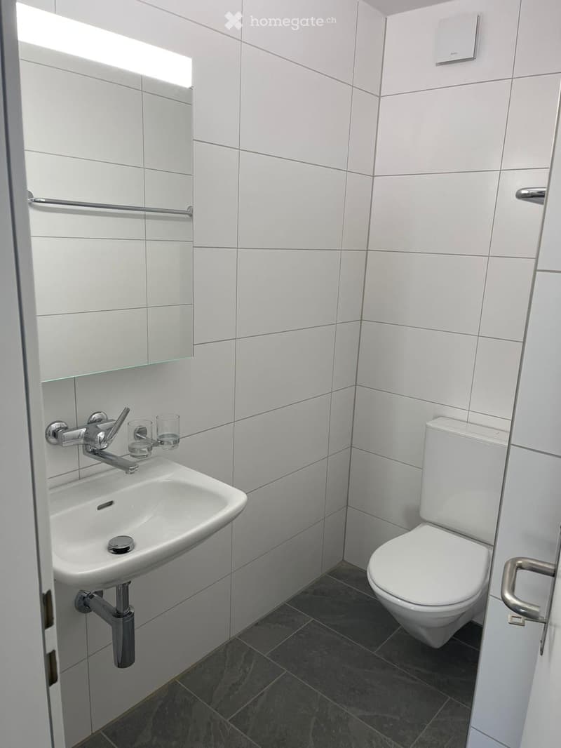 Modern bathroom with white tiled walls, a white sink with a chrome faucet, a toilet, and a mirror above the sink. Two small glass cups are placed on the sink counter.