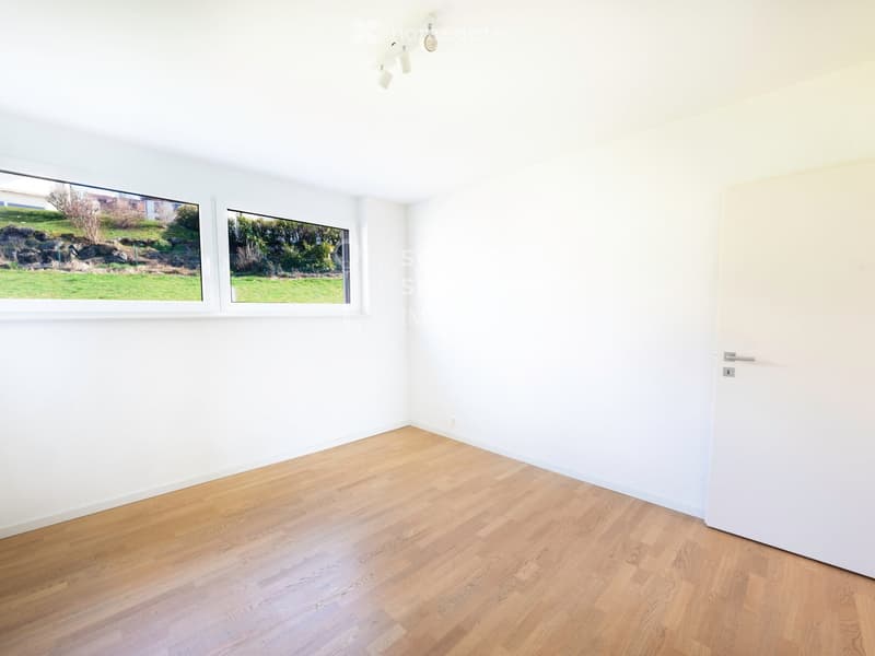 Empty room with light-colored wooden flooring and white walls, featuring large windows with a view of a garden and a door on the right side.