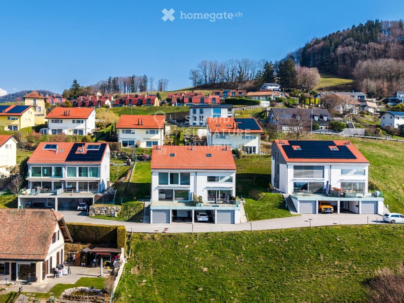 Aerial view of a residential neighborhood with multiple modern houses featuring red-tiled roofs, solar panels, and attached garages. The houses are surrounded by green lawns and trees, with a hilly landscape in the background.