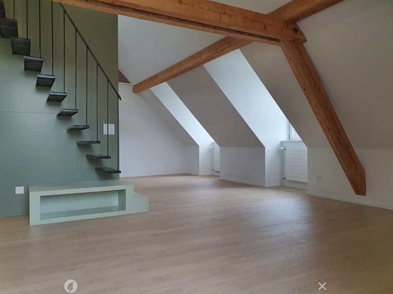 An empty attic space with wooden beams, wooden flooring, and a modern staircase leading upwards. The room has large skylights and white walls.