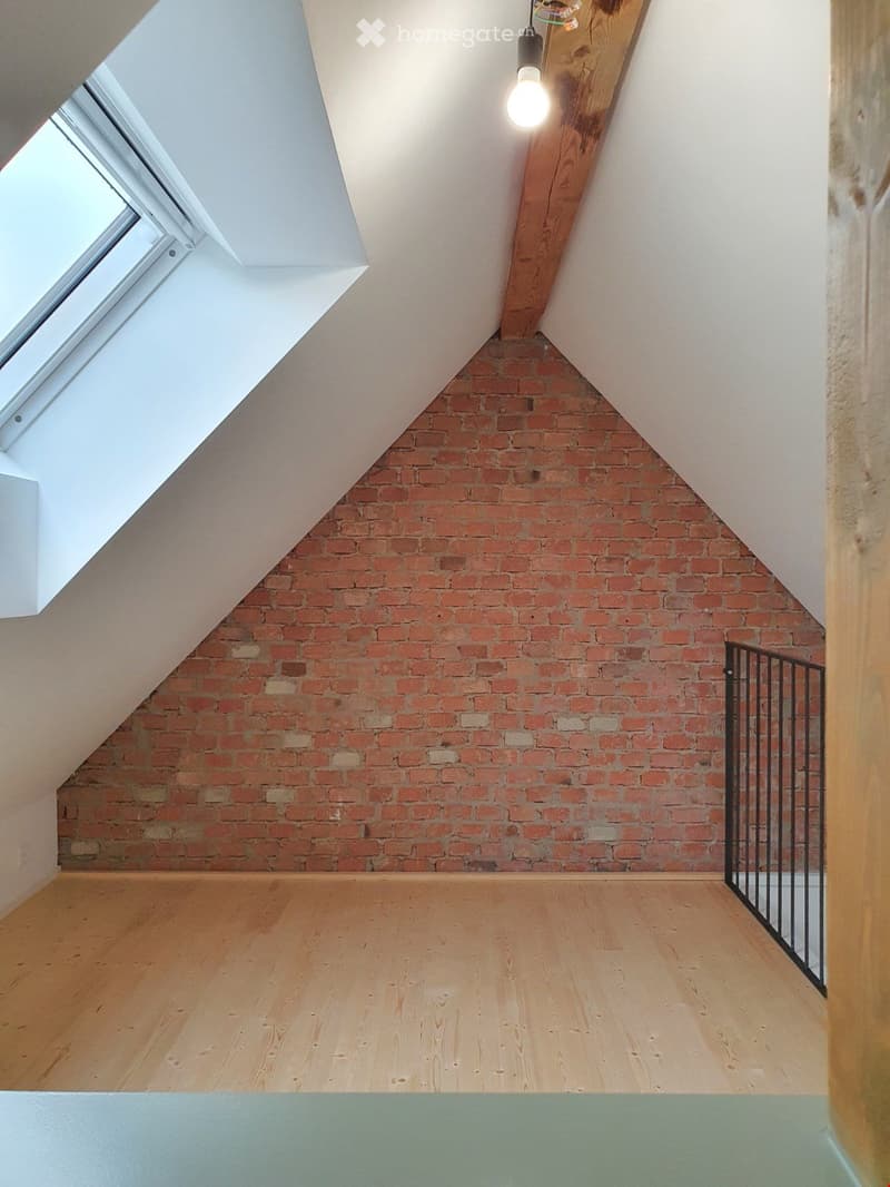 Empty attic space with exposed brick walls, wooden flooring, and a skylight window. The room features a simple, modern design with a hanging light fixture and a small black metal railing on one side.