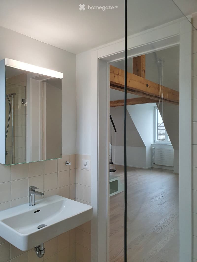 Modern bathroom with a white sink, chrome faucet, large mirror with built-in lighting, and a glass shower enclosure leading to a wooden loft area.