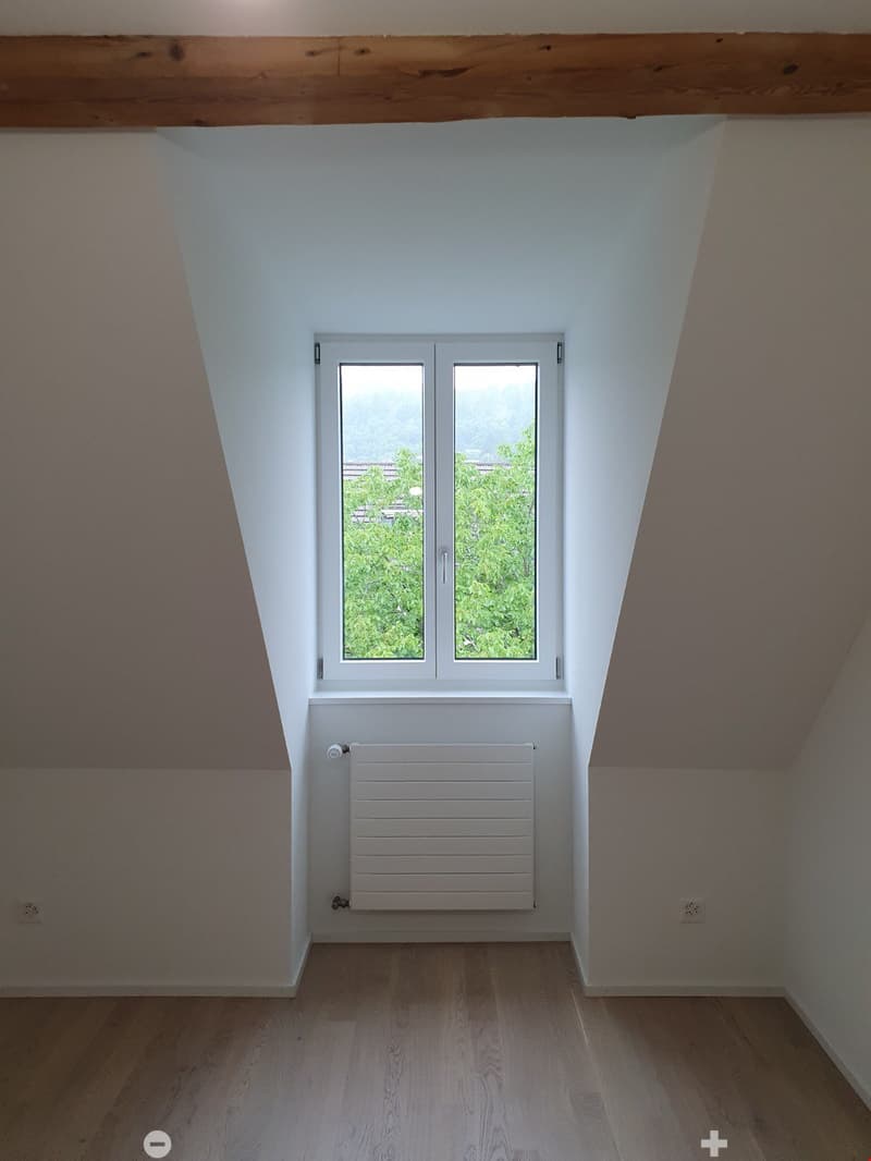 Empty attic room with wooden flooring, white walls, a large window offering a view of greenery, and a white radiator below the window.