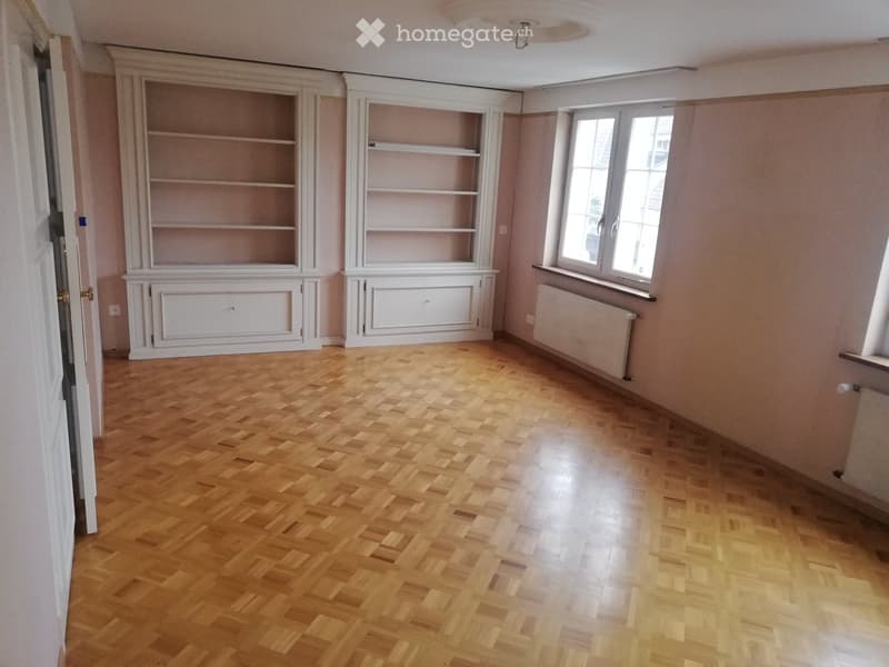 Empty room with wooden parquet flooring, built-in white shelving units, and large windows with radiators beneath them.