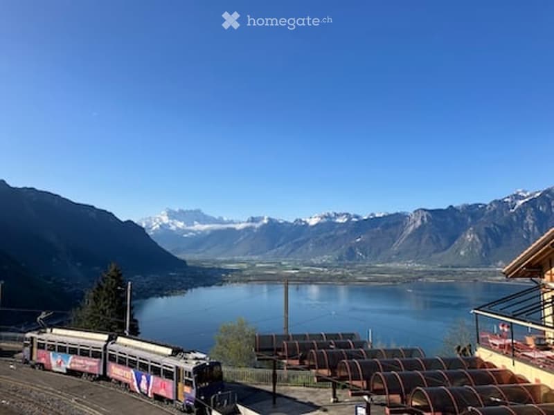 Scenic outdoor view of a lake surrounded by mountains, with a train passing by and a balcony overlooking the landscape.