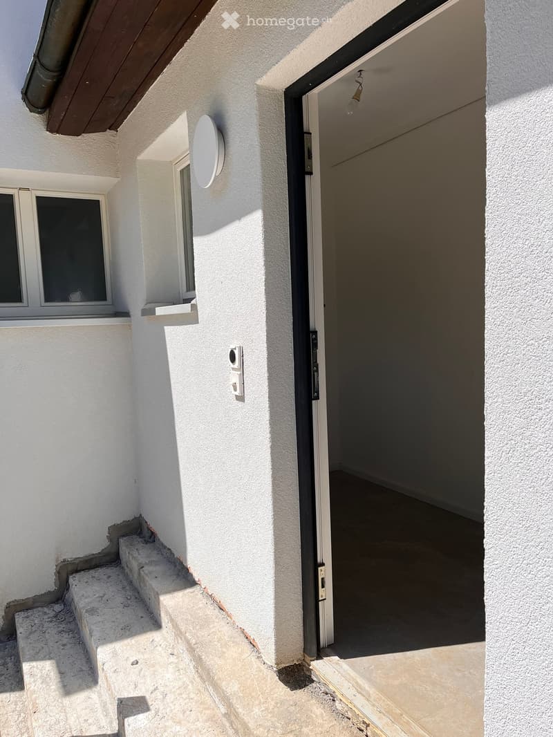 An exterior view of a building entrance with a raised ground floor, featuring a small staircase leading to a door, a window, and a satellite dish mounted on the wall.