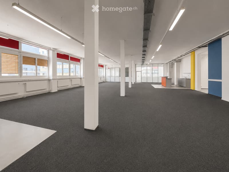 Open-plan office space with dark carpeted flooring, white walls, large windows with blinds, and minimal furniture including a small reception desk and a sink area.
