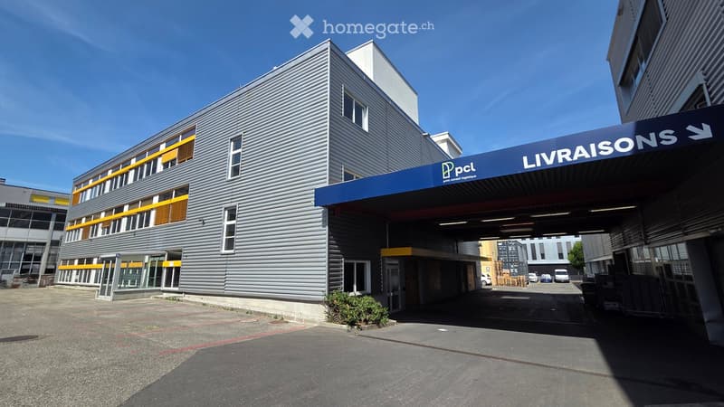 Modern industrial-style building with grey metal facade, large windows, and a blue sign reading "LIVRAISONS" above the entrance.