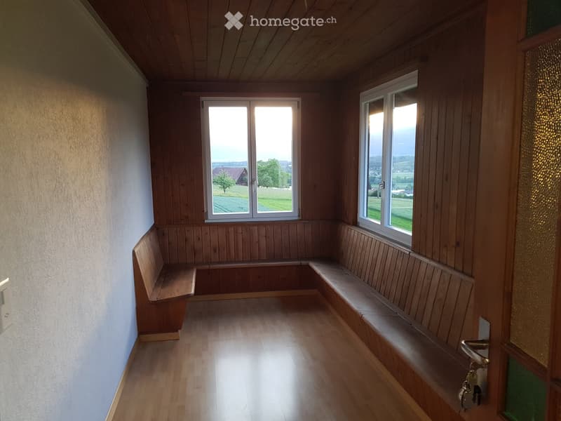 Empty room with wooden walls, wooden floor, built-in wooden benches along the walls, and large windows offering a view of the countryside.