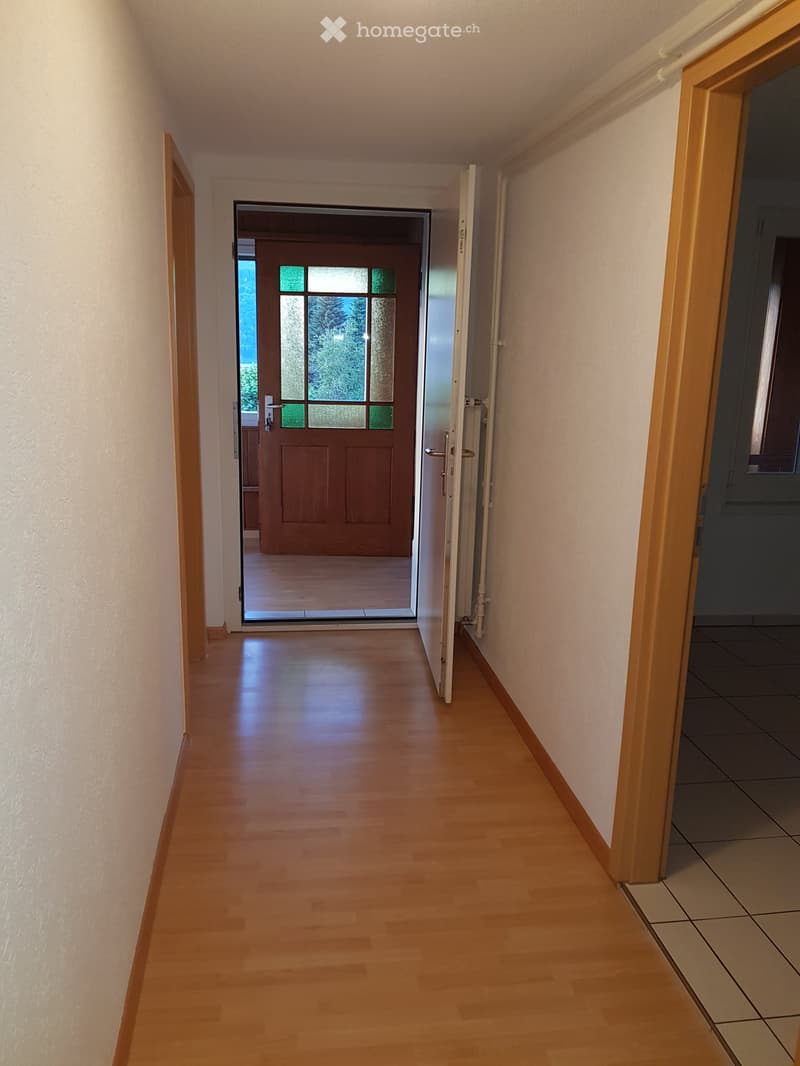 A clean, well-lit hallway with wooden flooring and white walls, featuring a partially open door leading to another room with a glass-paneled entrance door visible at the end.