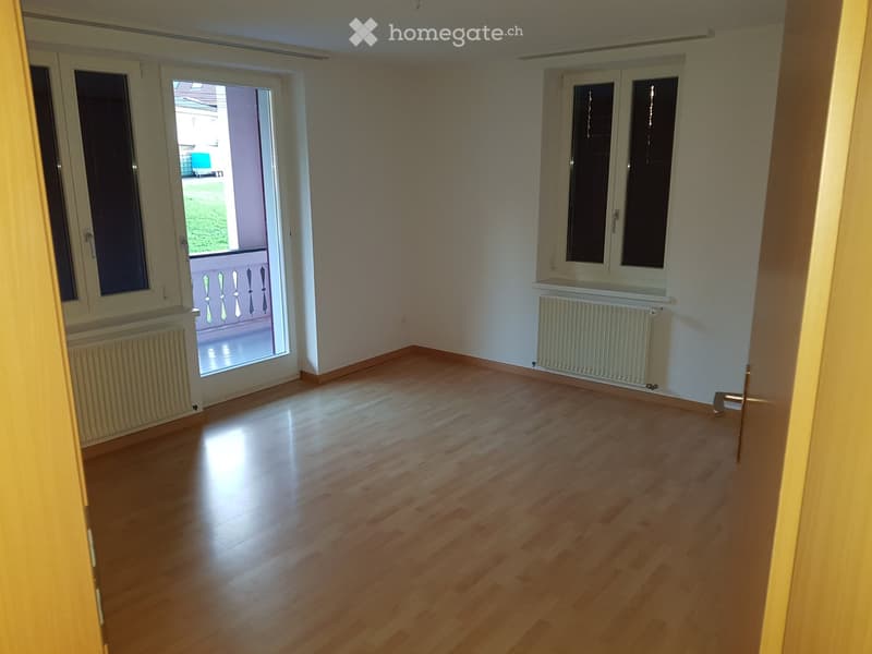 Empty room with wooden flooring, white walls, and large windows, including a sliding door leading to a balcony.