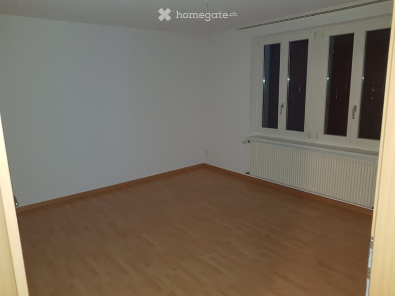 Empty room with light wooden flooring, white walls, and large windows with white frames. A radiator is visible beneath the windows.