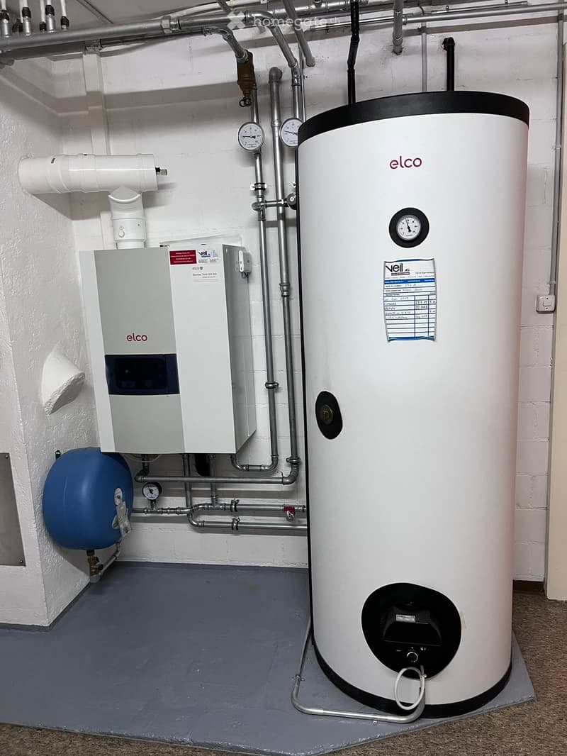 A utility room with a large white water heater, a pressure vessel, control unit, and various pipes and gauges.