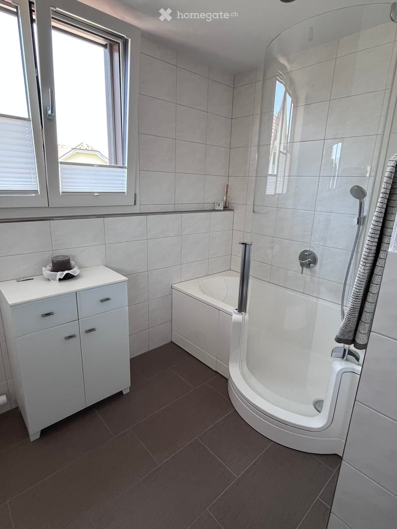 Modern bathroom with a bathtub, sink, and showerhead. The room features tiled walls and a window with blinds.