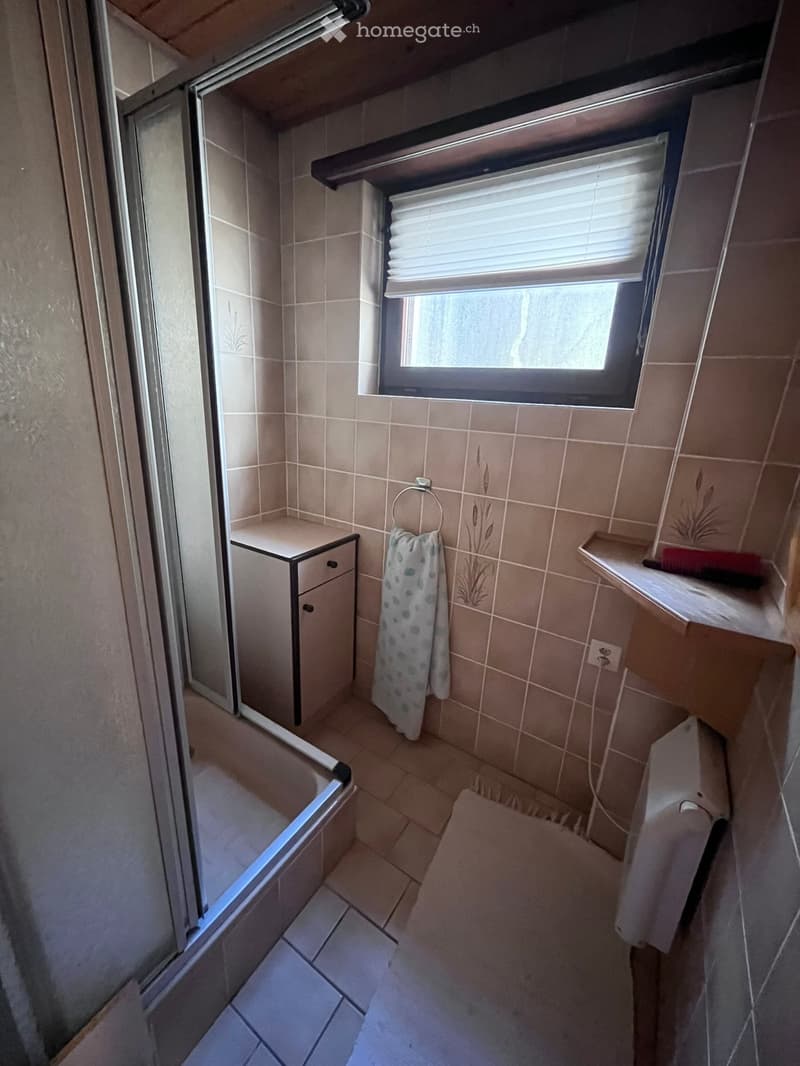 Small bathroom with tiled walls and floor, featuring a bathtub, a small cabinet, a towel, and a window with blinds.