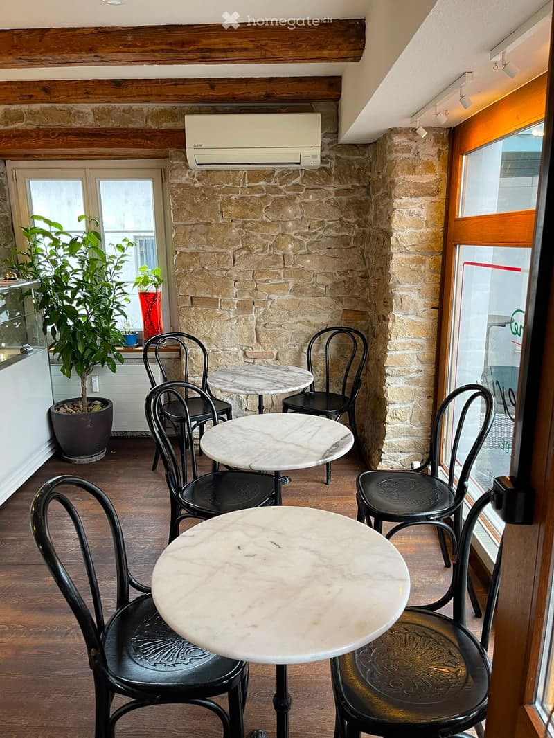A cozy, rustic-style room with exposed stone walls, wooden flooring, and large windows. It features several round marble tables and black metal chairs arranged for seating.