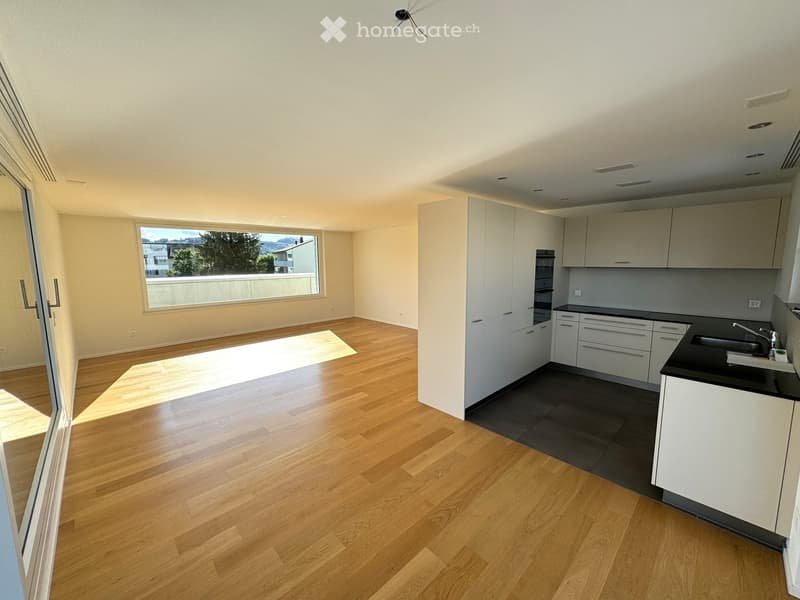 Modern kitchen with light wood flooring, white cabinets, and a black countertop. Features a large window with a view of trees and buildings.
