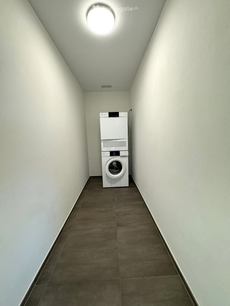 A narrow hallway with light grey walls and a brown tiled floor, featuring a white washing machine and dryer unit positioned against one wall.
