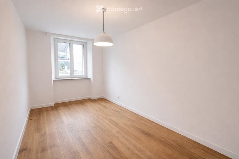 Empty room with light wooden flooring, white walls, and a single window with a view of the outside. A modern pendant light hangs from the ceiling.