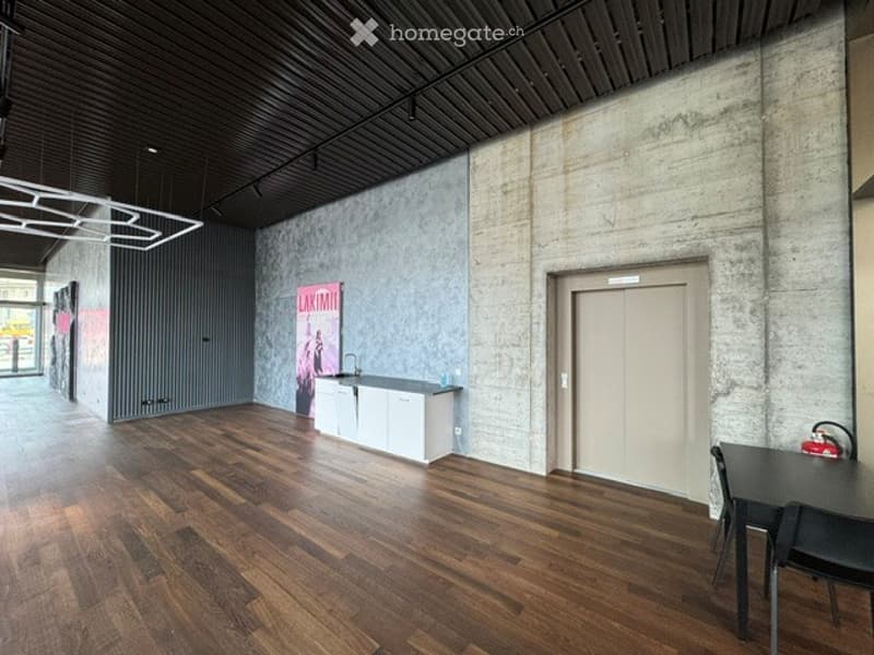 Modern, minimalist room with exposed concrete walls, polished wooden flooring, a small kitchen area with white cabinets and a sink, a dining table with chairs, and a red poster on the wall.