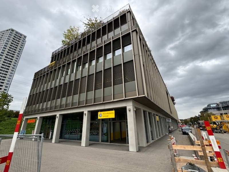 Modern multi-story building with a sleek, angular design and large glass windows. The entrance features a sign and a paved area with barriers.