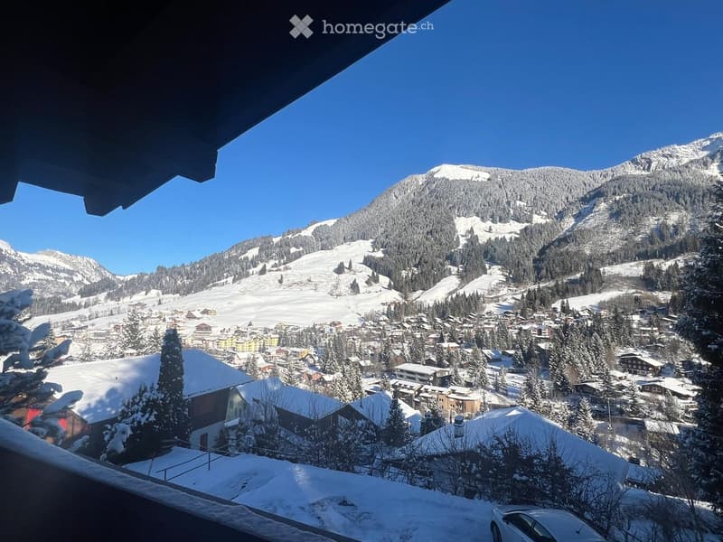 Panoramic view from a balcony overlooking a snow-covered village and mountain range under a clear blue sky.