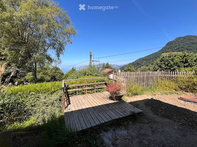A wooden deck with a railing, surrounded by greenery and a wooden fence, overlooking a scenic valley with mountains in the background.