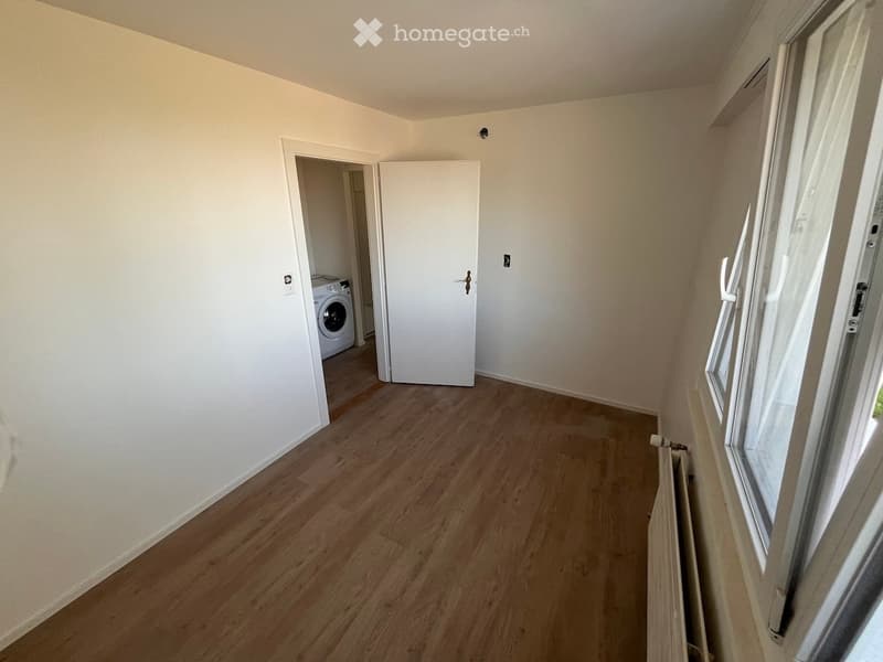 Empty room with light wooden flooring, white walls, and a door leading to a laundry area with a washing machine. A radiator is visible near the window.